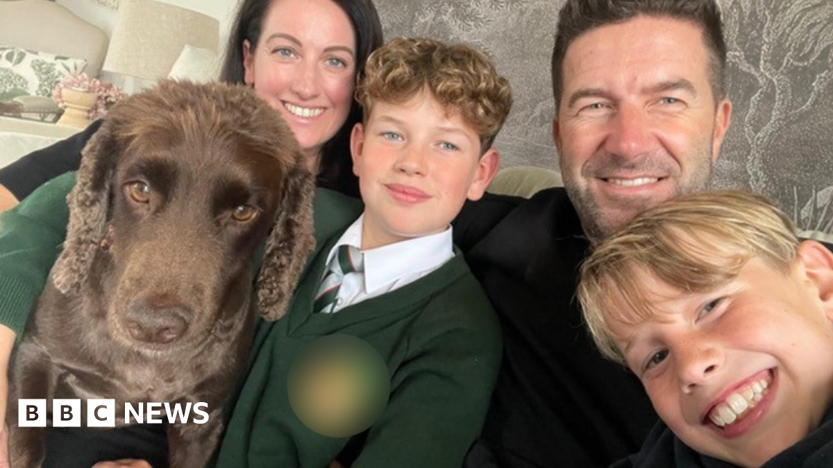 A woman and man, both with dark hair, sat on a sofa in front of a grey wallpaper backdrop. In front of them are two blonde boys, one with curly hair and the other with straight, and a brown cocker spaniel. All four people are looking at the camera and smiling.