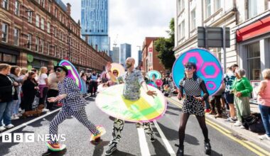 Three participants in a Manchester Pride parade. One wears a black trilby and black and white checked suit (left). The central marcher has a beard, black and white outfit with a multicoloured circle around the midriff while the person on the right has a multi patterned black and white dress  and dyed blue hair.