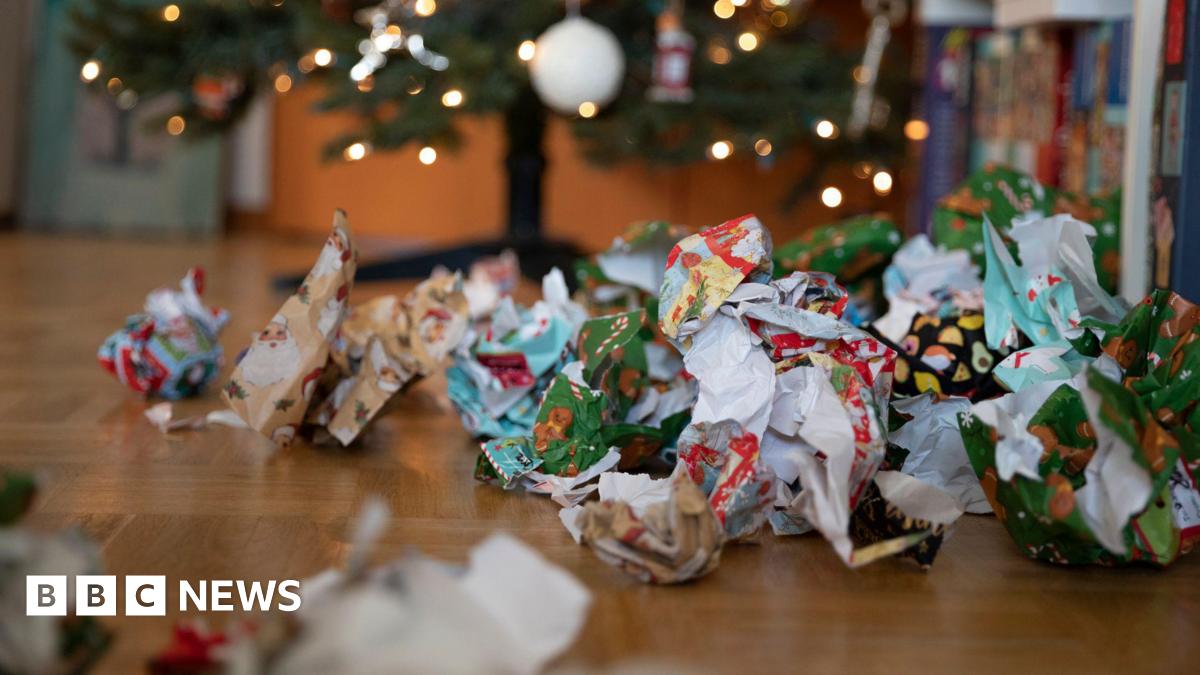 Colourful Christmas wrapping paper scrunched up in balls on a wooden floor in front of a decorated, lit up Christmas tree.