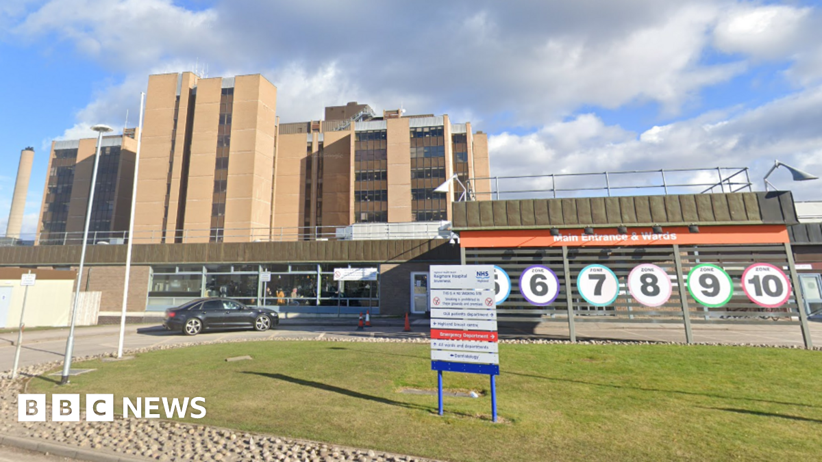 The main multi storey tower block at Raigmore Hospital is lit up by bright sunshine. A car has stopped next to the main entrance to the hospital. There are signs and numbers to help people find the right department.