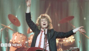 A man with a pink shirt and a red stripey tie on. He is also wearing a black suit jacket. He has long curly hair and is standing in the middle of a stage with a guitar.