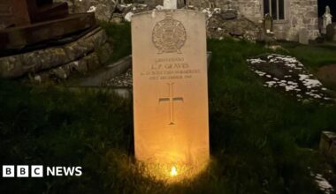 A row of three white gravestones arranged in a diagonal row across the shot.  It's night time so the area around them is in darkness.  Each has a small battery night light in front of it, so they are lit up in a warm orange glow.