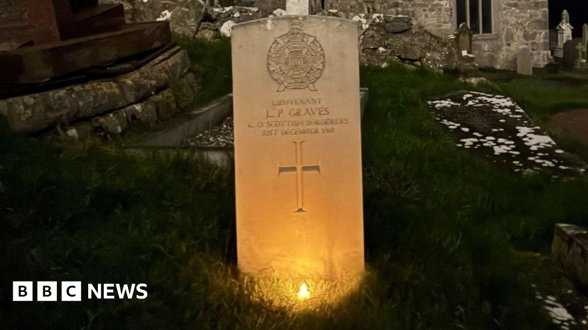 A row of three white gravestones arranged in a diagonal row across the shot.  It's night time so the area around them is in darkness.  Each has a small battery night light in front of it, so they are lit up in a warm orange glow.