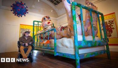 Artist Jason Wilsher-Mills sits alongside one of his exhibits - a giant inflatable character lying in a hospital bed watching TV inside the Grundy Art Gallery.