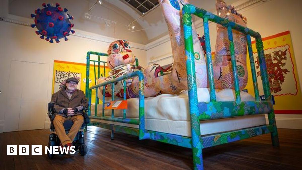 Artist Jason Wilsher-Mills sits alongside one of his exhibits - a giant inflatable character lying in a hospital bed watching TV inside the Grundy Art Gallery.