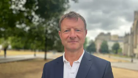 A man wearing a white shirt and jacket looking directly at the camera in front of a cathedral