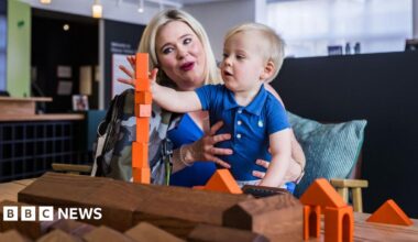 A woman with blonde hair sits at a table and holds a blonde haired toddler on her lap. The boy is stacking up orange building bricks in front of wooden models of buildings on the Elsecar Heritage Centre site.