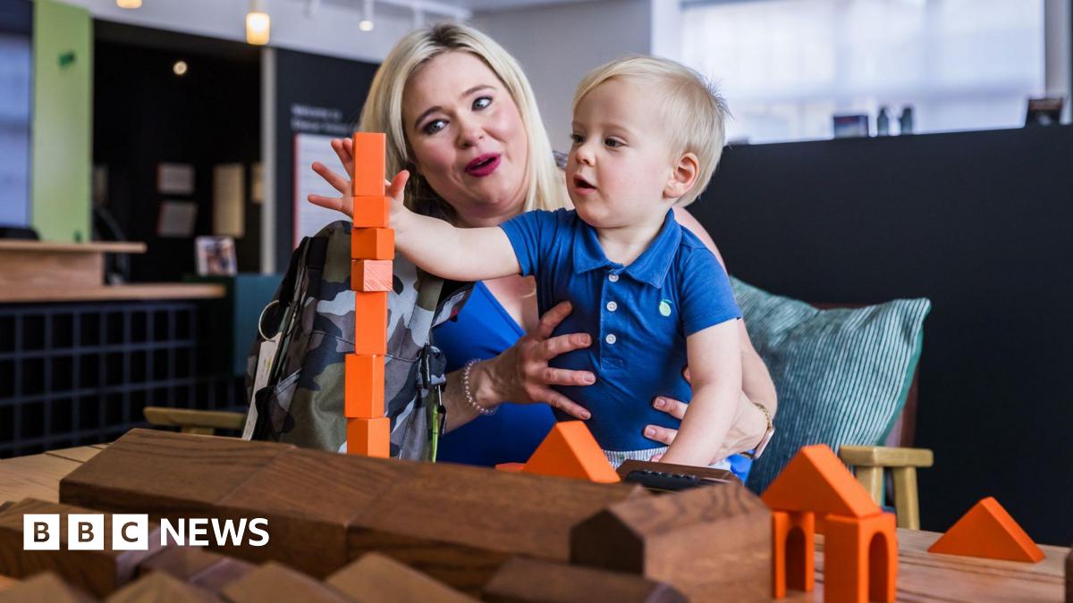 A woman with blonde hair sits at a table and holds a blonde haired toddler on her lap. The boy is stacking up orange building bricks in front of wooden models of buildings on the Elsecar Heritage Centre site.