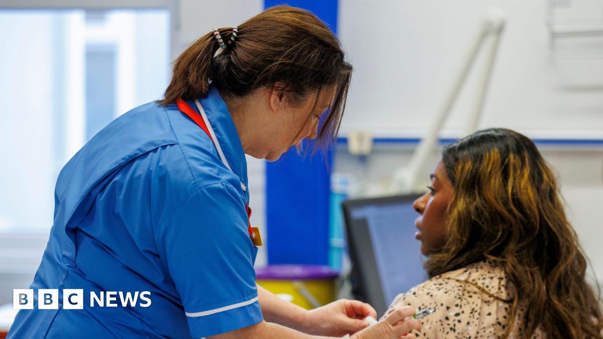 A nurse, dressed in uniform, is giving a vaccination to a patient. The nurse is standing to the left of the patient who is sitting down. She is wiping the patient's arm with cotton wool. They are inside a hospital.
