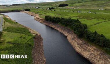 Aerial view of a reservoir with visibly low water levels, exposing wide, dry, rocky banks. Surrounding the reservoir are green fields, scattered trees, and a road running parallel to the water.
