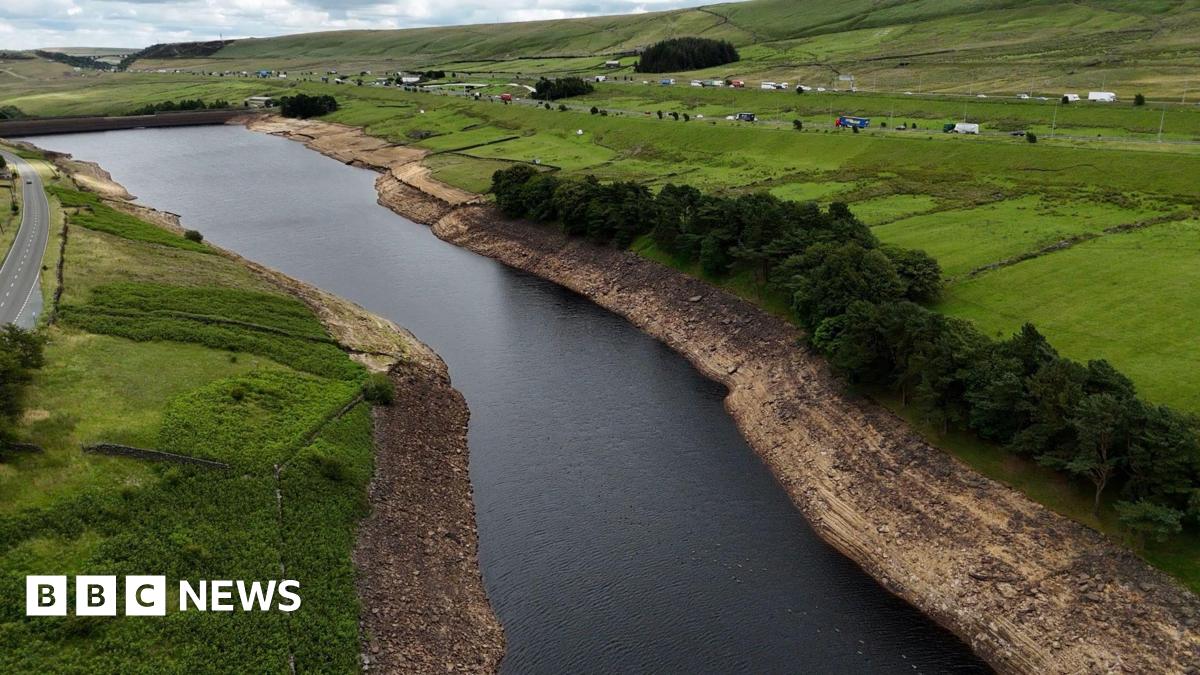 Aerial view of a reservoir with visibly low water levels, exposing wide, dry, rocky banks. Surrounding the reservoir are green fields, scattered trees, and a road running parallel to the water.