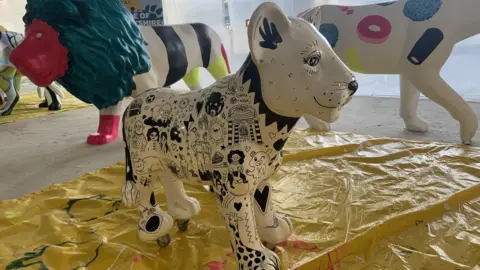 A small statute of a white lion cub is painted white with a black design of cartoons including animals and faces