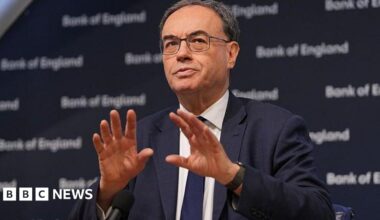 A lifeguard runs down a road barefoot carrying a red defribrillator bag next to parked cars on a road with trees and the sea in the background.