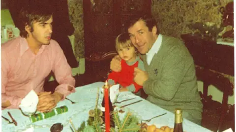 Rob Parsons Photograph of Ronnie Lockwood (right) holding onto Rob and Dianne's young son. He sits at the dining table with Rob (left) who wears a pink shirt. Candles and wine bottles can be seen on the table, along with cutlery and Christmas crackers. 
