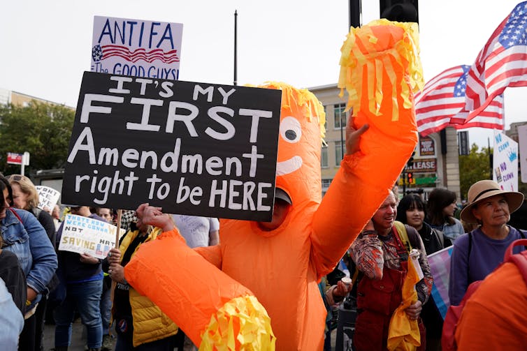 A protest with a person in an orange outfit carrying a sign saying 'It's my First Amendment right to be HERE.'