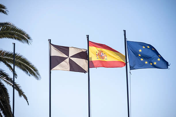 Three flags—black and white, Spain, and the EU—flying on tall poles against a clear blue sky.