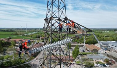Apprentices wearing safety harnesses and orange vests work on top of a high pylon, practising maintenance on overhead power lines