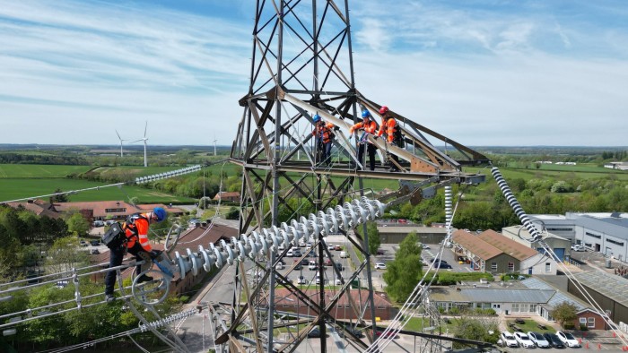 Apprentices wearing safety harnesses and orange vests work on top of a high pylon, practising maintenance on overhead power lines