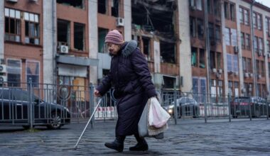 An elderly woman with a cane walks past a building in Kyiv with shattered windows and visible structural damage, caused by a Russian strike.