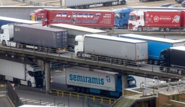 Multiple large trucks and lorries travel on elevated and ground-level roads at the Port of Dover, with shipping containers and company logos visible.