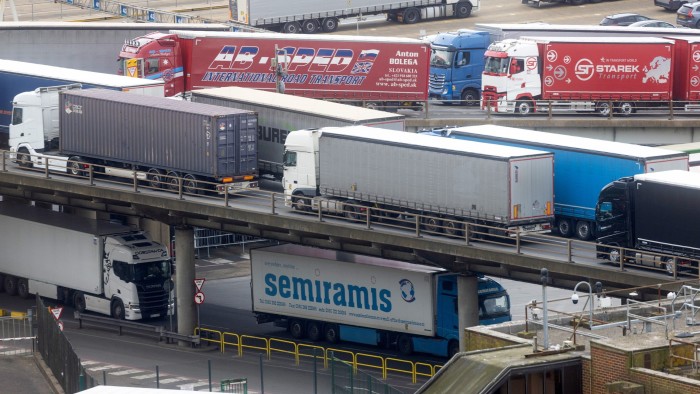 Multiple large trucks and lorries travel on elevated and ground-level roads at the Port of Dover, with shipping containers and company logos visible.