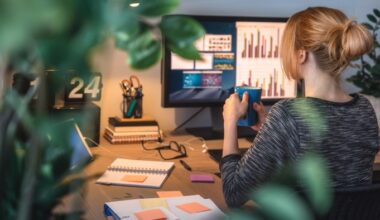 A woman sits at a desk with a computer displaying charts, holding a mug