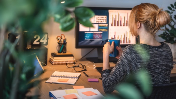 A woman sits at a desk with a computer displaying charts, holding a mug