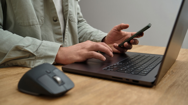 Closeup image of a man using a laptop and phone on a wooden desk table.
