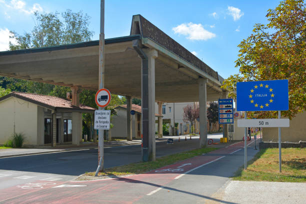 A roadside border crossing with an EU “Italia” sign stands near an old checkpoint structure on a sunny day.