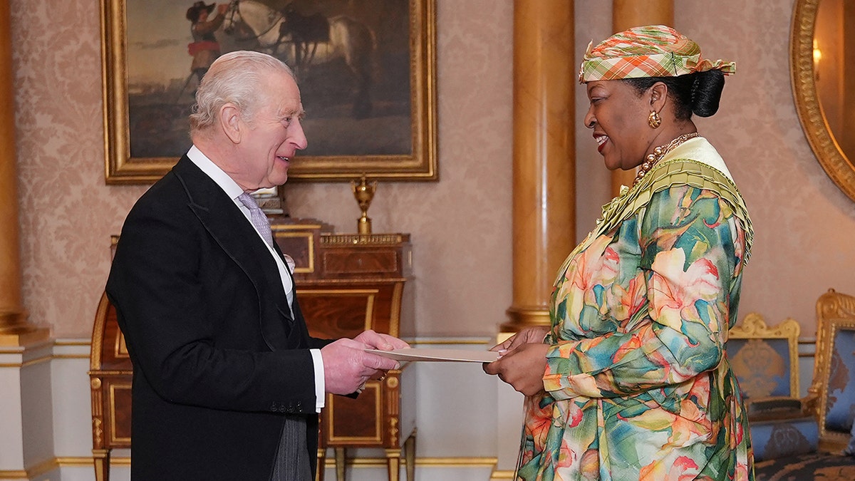 King Charles smiling at a woman as they converse inside Buckingham Palace during a royal visit.
