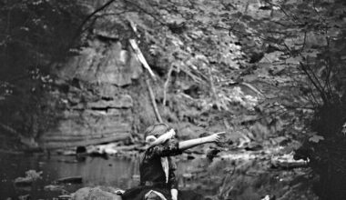 A young girl with a blindfold points to the right in front of a stream