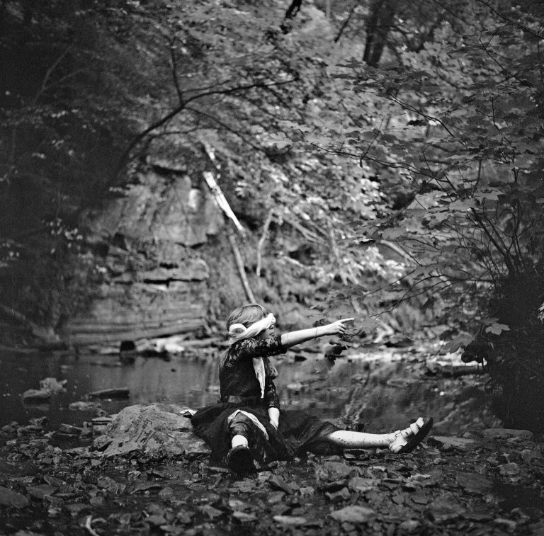 A young girl with a blindfold points to the right in front of a stream