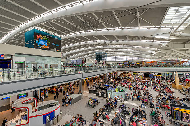 A wide view of a busy airport terminal with crowds of passengers, seating areas, and shops under a large curved roof.