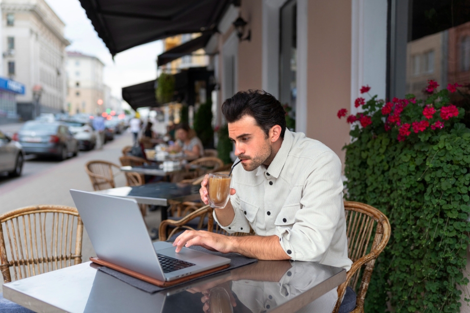 Photograph of a man working on a laptop at an outdoor café while drinking iced coffee.