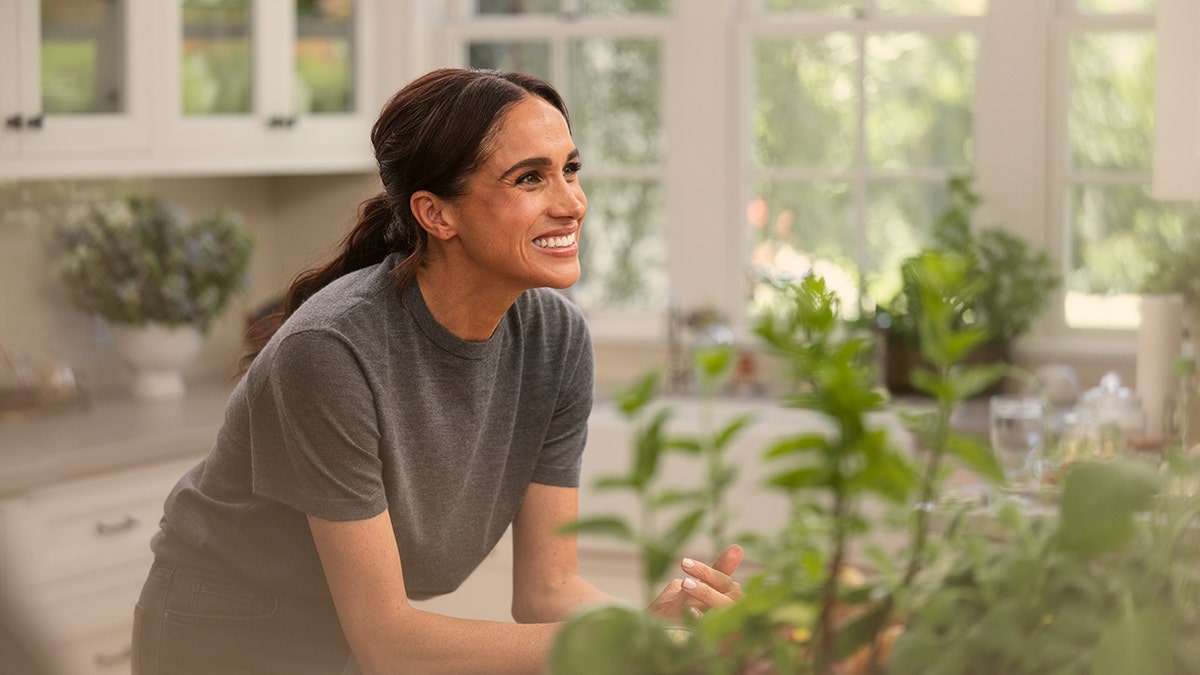 Meghan Markle looking up smiling wearing a grey sweater in a kitchen.