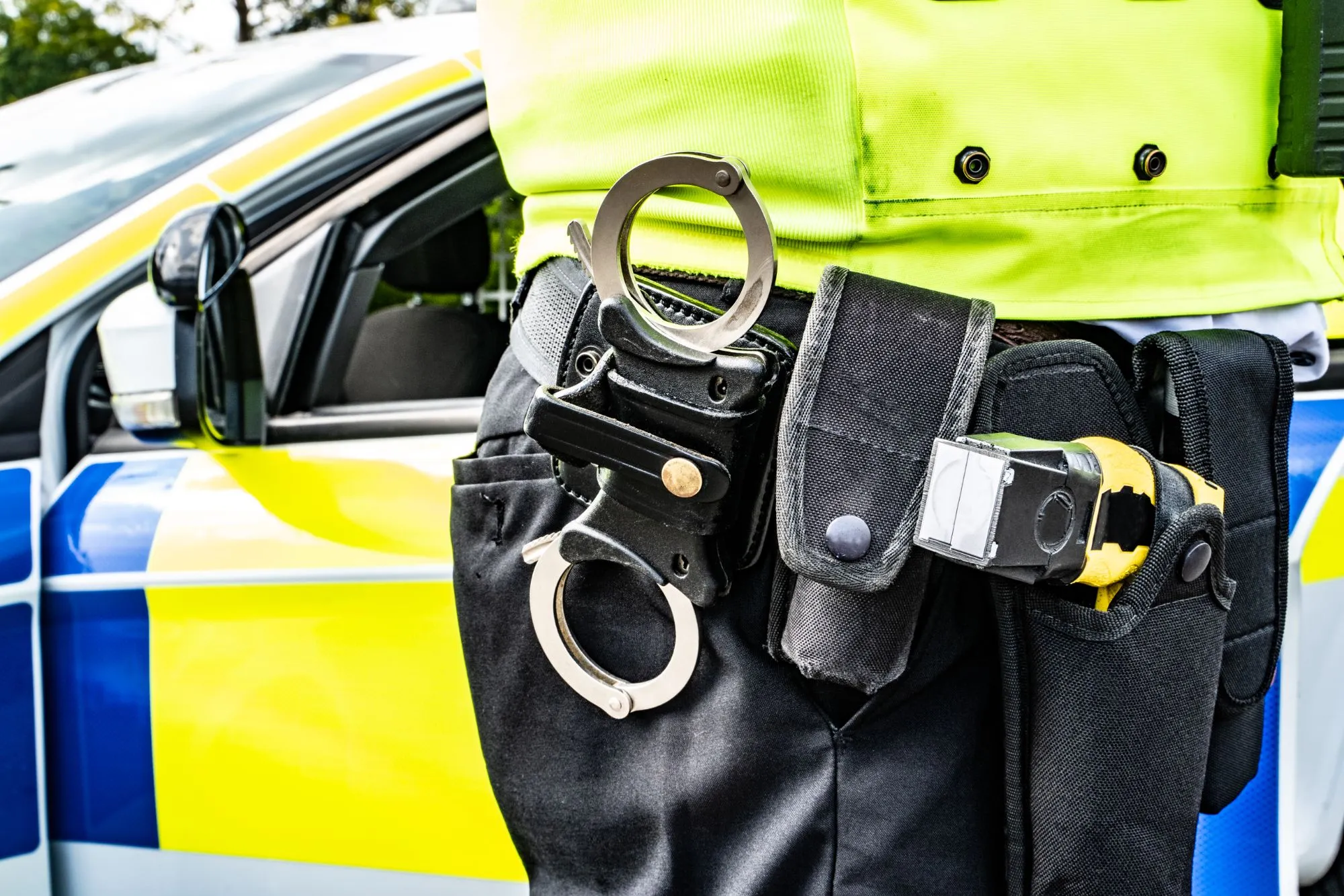 An image collage containing 1 images, Image 1 shows Close-up of a police officer's handcuffs on their equipment belt, with a police car in the background