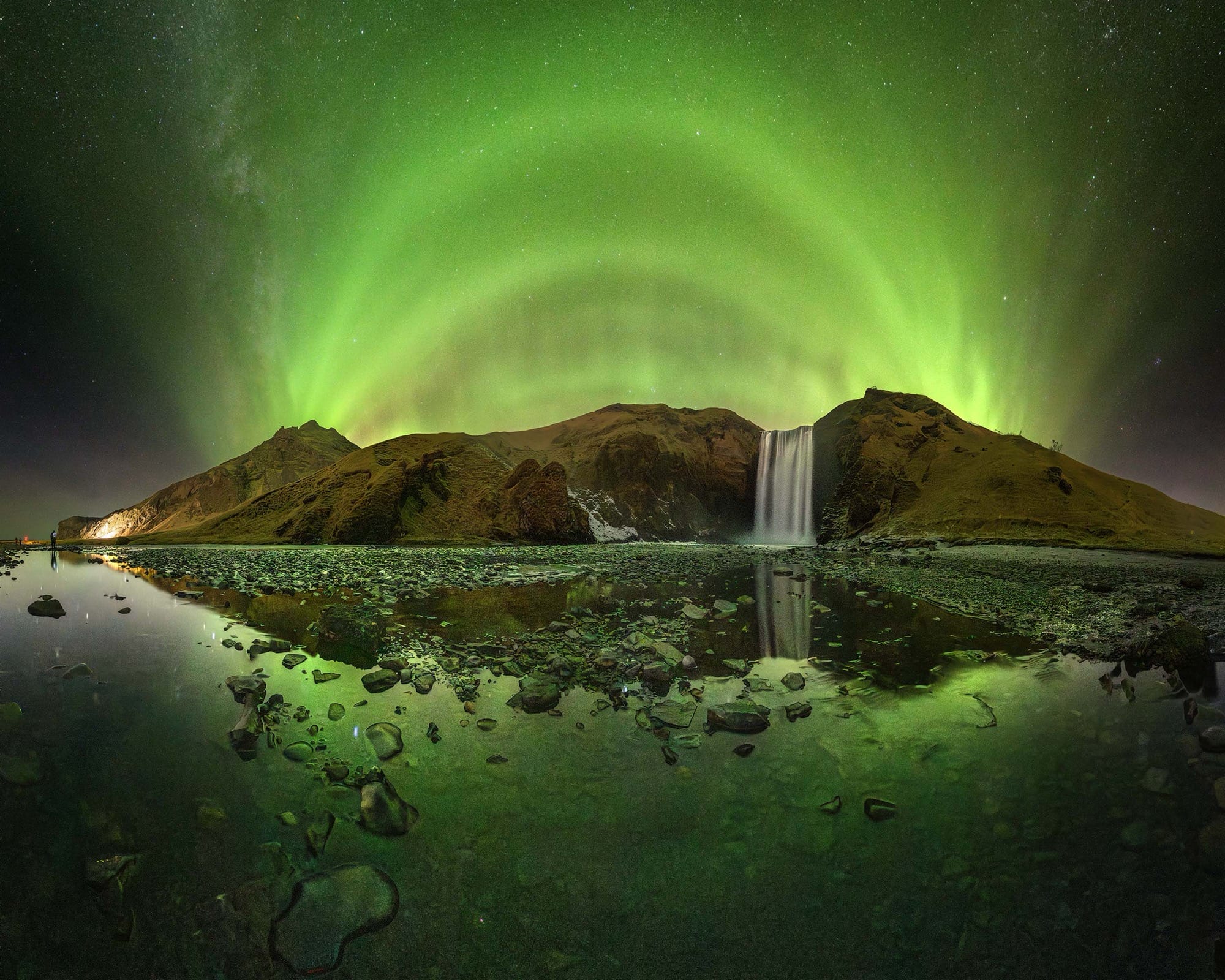 Arched green aurora borealis over a rocky landscape in Iceland, with a waterfall