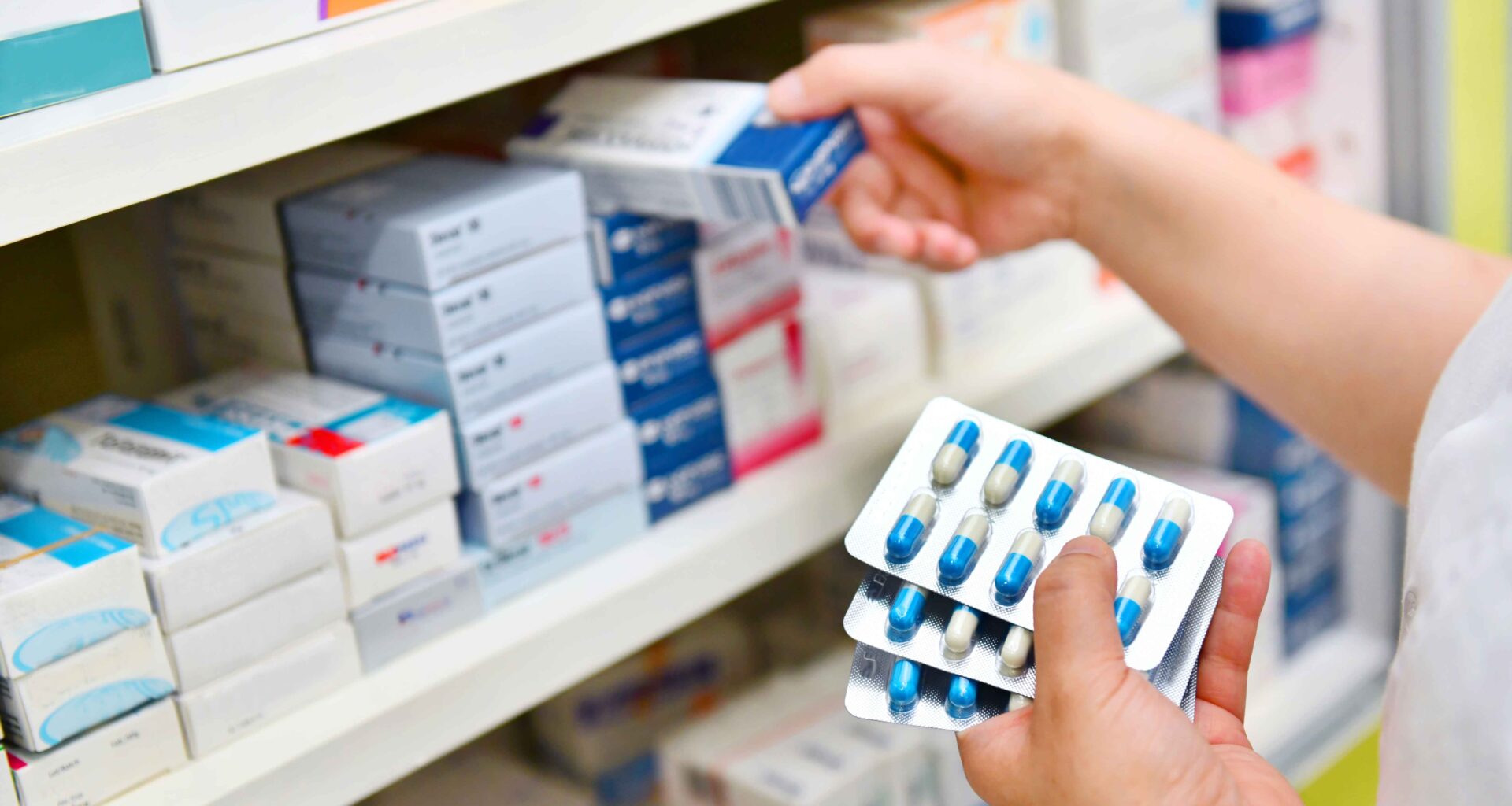 Person selecting medicine boxes from a pharmacy shelf while holding blister packs of capsules.