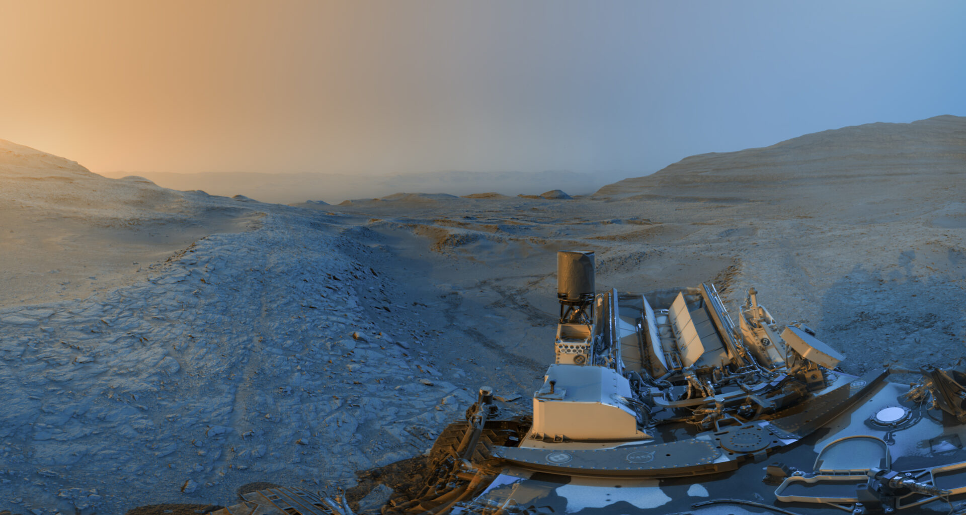 The Curiosity rover is visible at the bottom right of this panorama image. The rover is at the top of a ridge referred to as a boxwork formation. The sky ahead of it is hazy; it's red on the left and blue on the right.