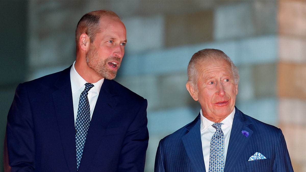 Prince William stands next to King Charles wearing a blue suit.