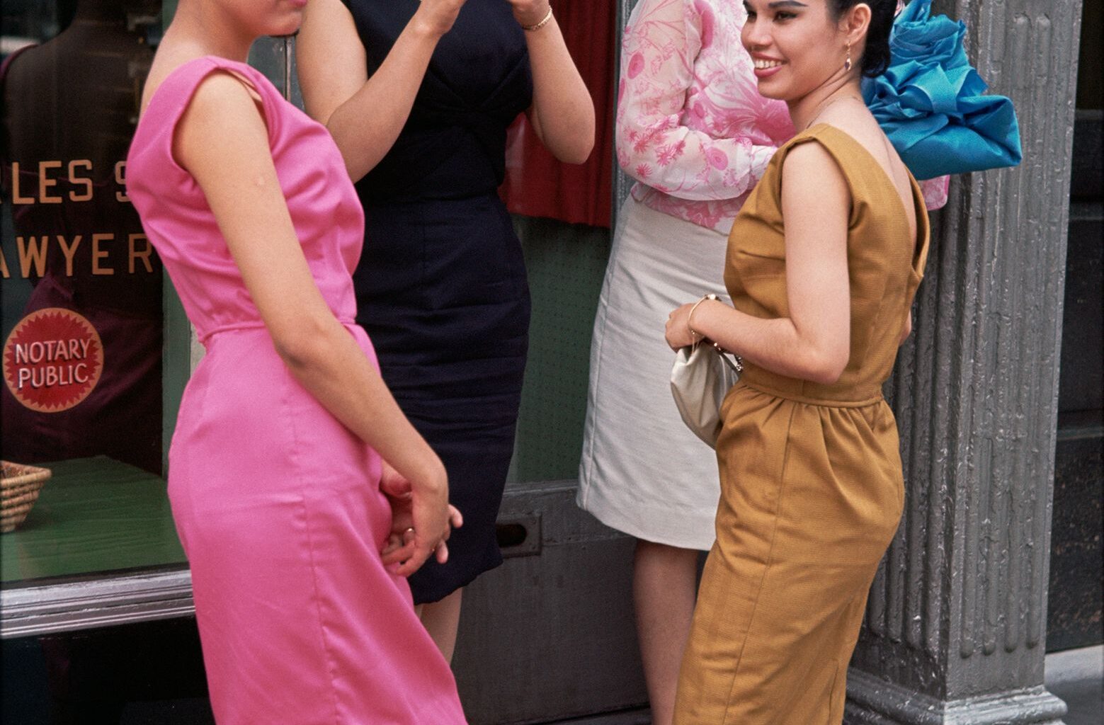 A group of four women in colorful 1960s dresses stand on a sidewalk, engaged in conversation outside a storefront