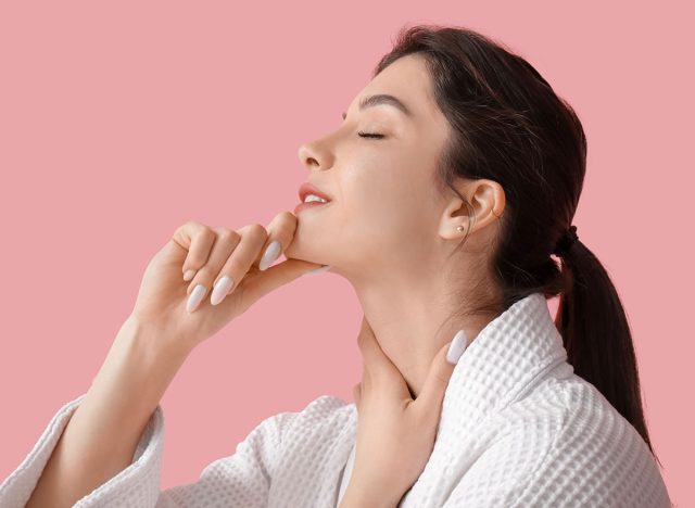 Beautiful young woman in bathrobe doing face building exercise on pink background