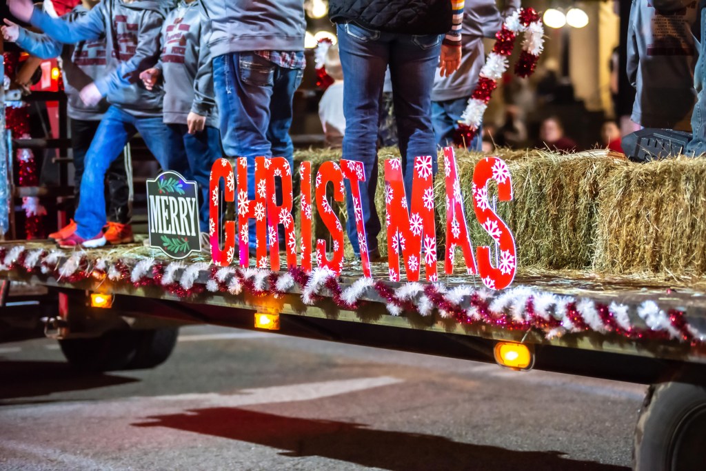 A Christmas parade float with red "Christmas" letters, a "Merry" sign, hay bales, and people in the background.