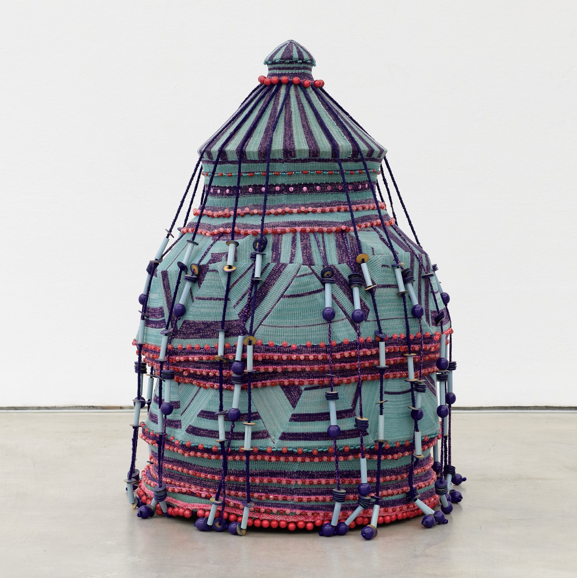 A woven textile sculpture by Jim Drain loosely resembling a large teal and magenta basket with a lid and many long strands or tassels