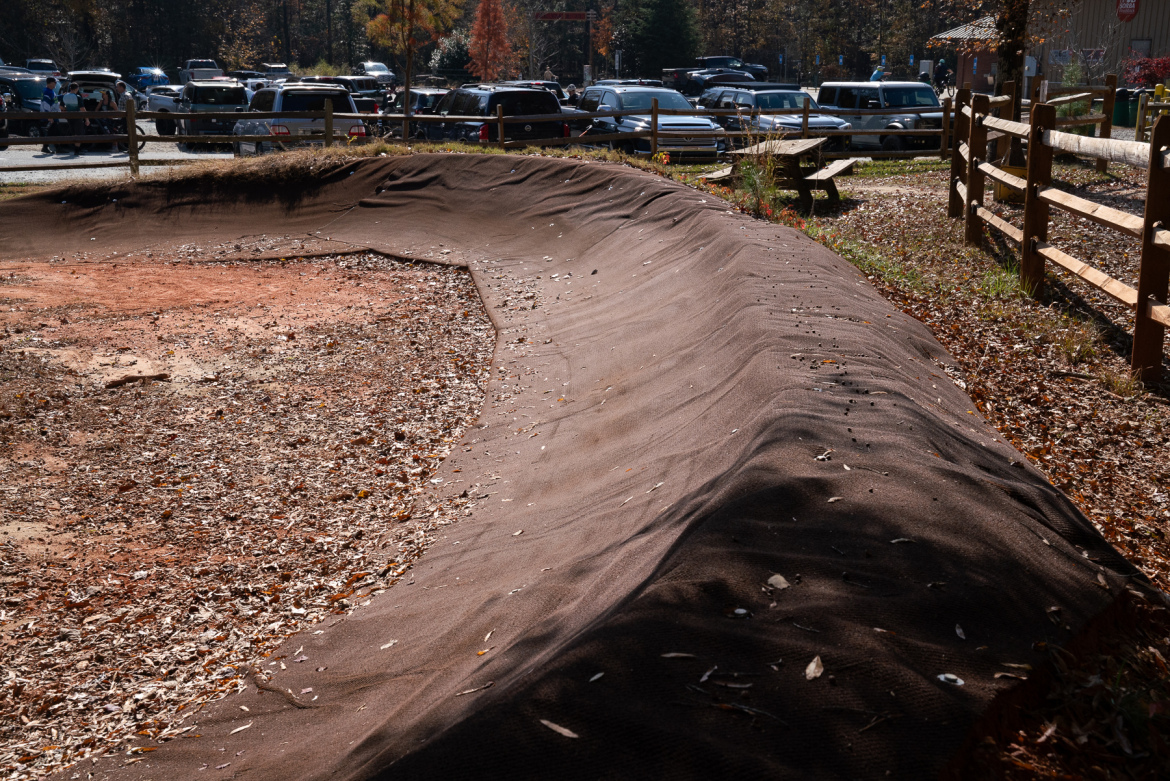 A winding dirt path with a curved edge, surrounded by fallen leaves, leading to a parking area with several vehicles in the background. A wooden fence runs alongside the path, while picnic tables are visible nearby, set against a backdrop of colorful autumn trees.