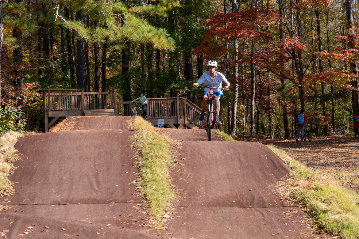 A young cyclist performing a wheelie on a dirt bike track surrounded by autumn foliage. In the background, another cyclist is seen on a wooden platform, and a few people are standing nearby.