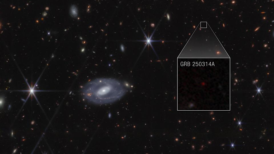 Webb image shows hundreds of galaxies of all shapes and sizes against the black background of space. Toward the center-left is a large white spiral galaxy that is almost face-on. To the right of this is a large box, which zooms in on an area at top right. Within the box is a faint red dot at the center, with the label GRB 250314A.