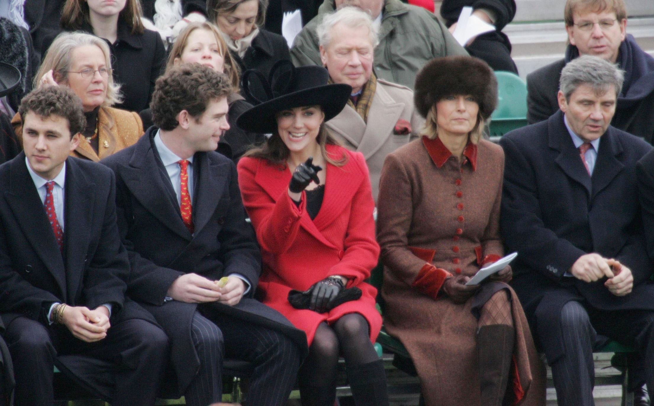 Kate Middleton wearing a red coat and pointing, sitting next to her parents and friends at William's graduation