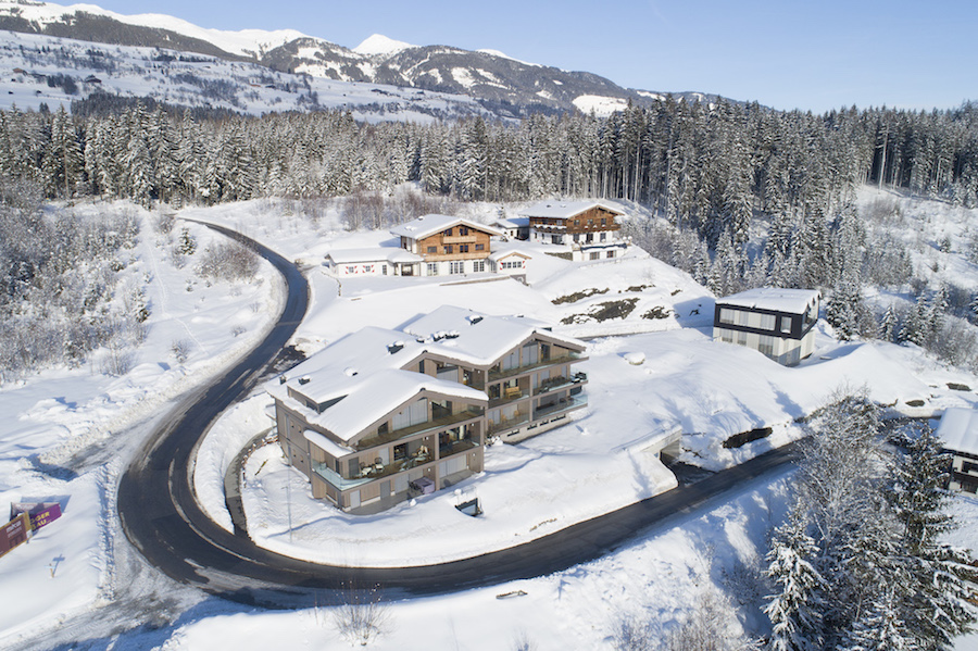 Aerial view of multiple snow-covered buildings in Hollersbach, Austria, on a winding road with a forest and mountains in the background.