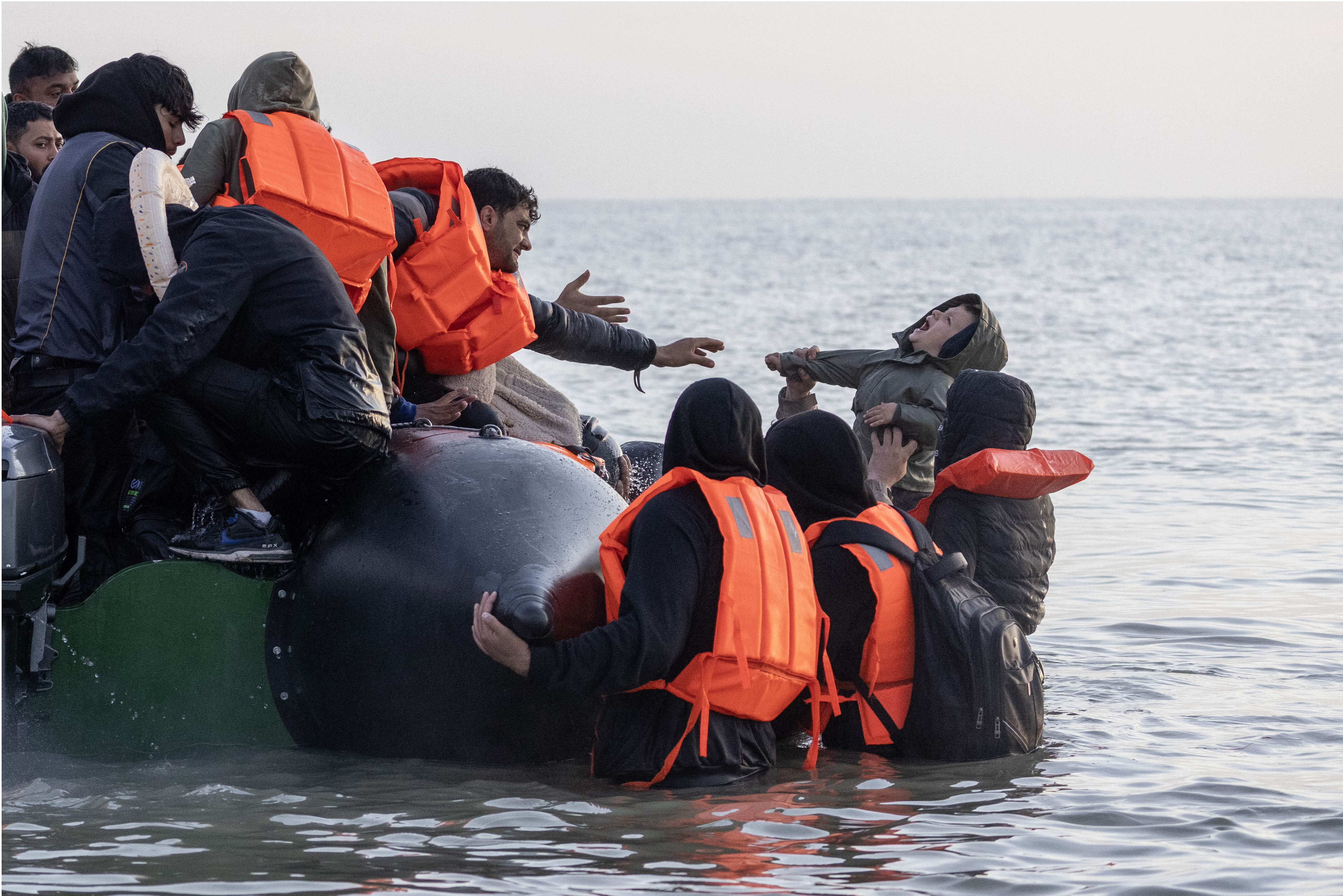 Migrants in orange life jackets prepare to cross the English Channel in a boat.